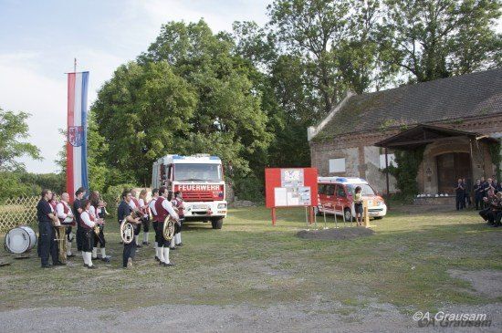 Eine Gruppe von Menschen in traditionellen Uniformen spielt mit Blechblasinstrumenten vor einem Feuerwehrauto und einem Van. Dahinter ist eine Fahne auf einem Mast befestigt.