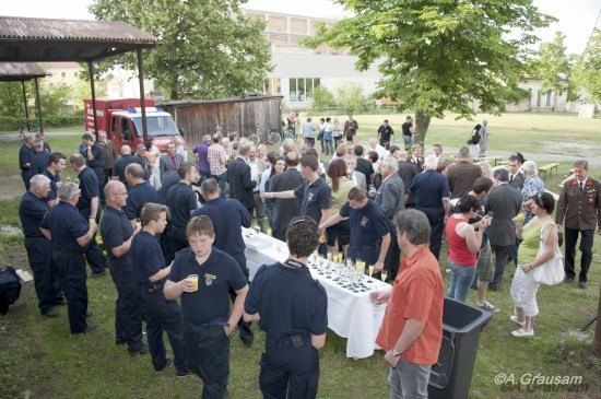 Eine große Gruppe von Menschen, einschließlich Feuerwehrleute in Uniform, versammelt sich um einen Tisch mit Getränken und Essen in einem Park. Ein Feuerwehrauto ist in der Nähe geparkt.