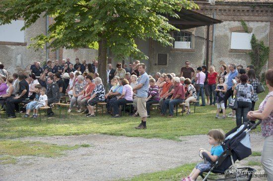 Eine Gruppe von Menschen sitzt in Reihen unter einem Baum, mit einem Mann vor ihnen. Einige tragen Sonnenbrillen, und eine Frau mit einem Kinderwagen ist in der Nähe. Die Umgebung ist im Freien, mit einem Gebäude im Hintergrund.