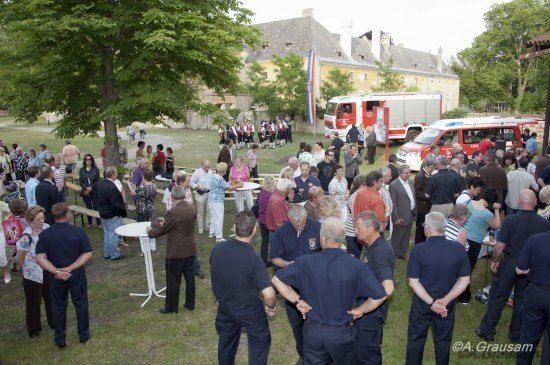 Eine große Gruppe von Menschen versammelt sich in einem Park mit Feuerwehrautos und einem Gebäude im Hintergrund. Einige tragen Uniformen, andere sind in Freizeitkleidung.