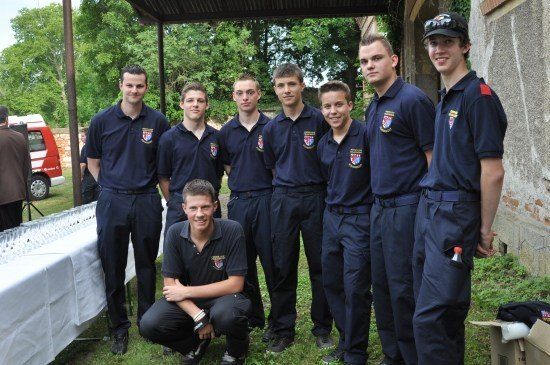 Eine Gruppe junger Männer in blauen Uniformen, einer kniet und andere stehen, posiert für ein Foto in einem Grasbereich mit einem Tisch und einem roten Fahrzeug im Hintergrund.