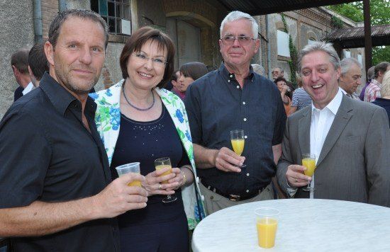 Vier Personen, drei Männer und eine Frau, stehen zusammen, lächeln und halten Gläser mit Orangensaft. Sie befinden sich bei einer Veranstaltung im Freien.