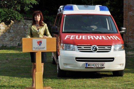 Eine Frau steht an einem Podium mit einem Mikrofon vor einem roten und weißen Feuerwehrauto mit dem Wort 'Feuerwehr' auf der Front.