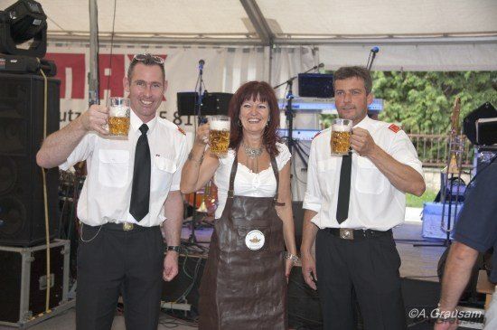 Drei Menschen in formeller Kleidung, zwei Männer und eine Frau, halten Gläser mit Bier in einer festlichen Umgebung mit Musikinstrumenten und Lautsprechern im Hintergrund.