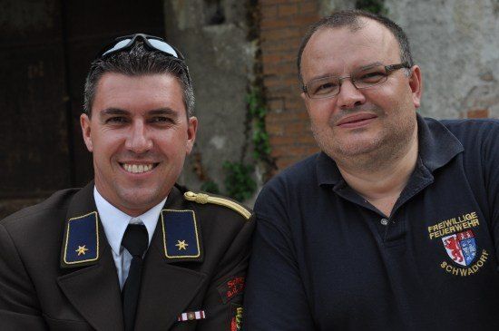 Zwei lächelnde Männer, einer in Uniform mit einem Stern auf der Schulter und der andere in einem marineblauen Poloshirt mit einem Logo auf der linken Brust.
