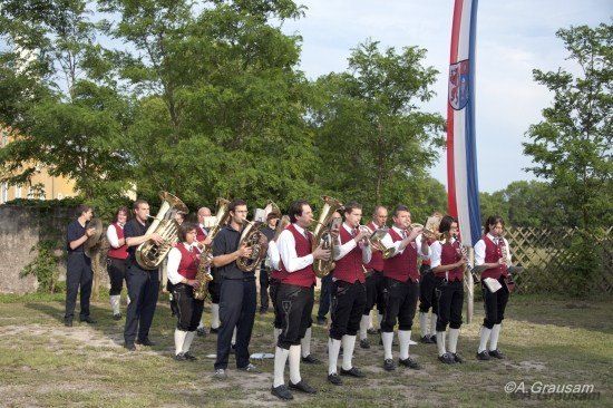 Eine Brassband in traditioneller Kleidung steht in einer Reihe auf einem Rasenfeld, mit Bäumen und einem Fahnenmast im Hintergrund.