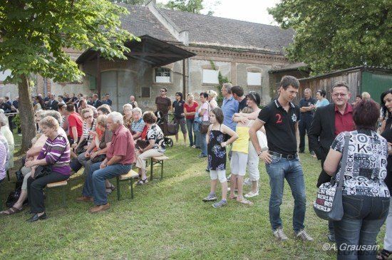 Eine Gruppe von Menschen versammelt sich im Freien, einige sitzen auf Bänken und andere stehen, mit einem Gebäude im Hintergrund.