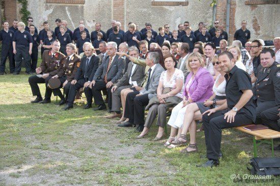 Eine große Gruppe von Menschen in formeller Kleidung sitzt auf Bänken vor einem Backsteingebäude. Einige tragen Uniformen, andere sind in Anzügen und Kleidern. Sie scheinen an einer Veranstaltung teilzunehmen.