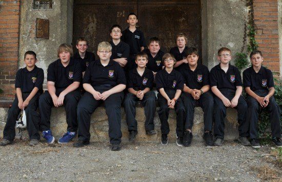 Eine Gruppe junger Jungen in passenden schwarzen Uniformen sitzt auf einer Steinbank vor einem Ziegelgebäude. Sie alle lächeln und scheinen für ein Foto zu posieren.