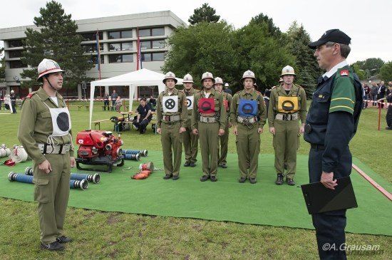 Eine Gruppe uniformierter Personen, möglicherweise Feuerwehrleute, stehen auf einem grünen Feld mit einem Mann in schwarzer Uniform, der eine Mappe hält. Im Hintergrund befinden sich Ausrüstungsgegenstände und ein Zelt.