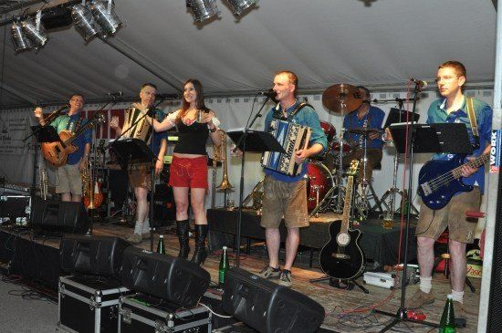 Eine Band spielt auf einer Bühne mit verschiedenen Instrumenten, darunter Akkordeon, Gitarre und Schlagzeug, unter einem weißen Zelt.