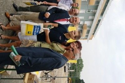Eine Gruppe von Menschen in formeller Kleidung steht lächelnd vor einem Gebäude und posiert für ein Foto. Einige halten eine weiße Tasche und eine grüne Mappe.
