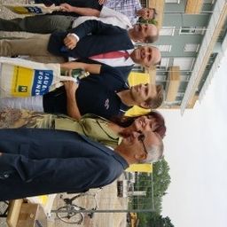 Eine Gruppe von Menschen in formeller Kleidung steht lächelnd vor einem Gebäude und posiert für ein Foto. Einige halten eine weiße Tasche und eine grüne Mappe.