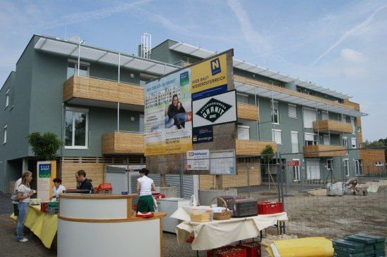 Eine Gruppe von Menschen steht vor einem Gebäude im Bau. Es gibt Tische mit gelben Tüchern, einem Korb und Lagerboxen. Ein Schild mit einem Bild eines Paares ist an der Wand montiert.