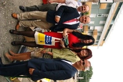 Eine Gruppe von Menschen steht zusammen draußen und lächelt für ein Foto. Eine Frau in einem roten Kleid hält eine gelbe Tasche mit dem Wort 'Home' darauf. Hinter ihnen steht ein Gebäude mit einem Schild.