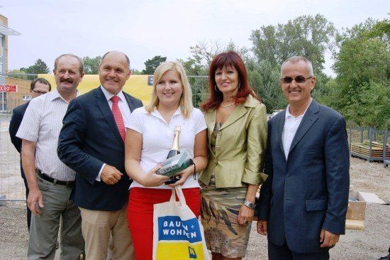 Eine Gruppe von Menschen posiert für ein Foto mit einer Frau, die eine Flasche Champagner und eine Tasche mit dem Text Baun+Wohnen hält.