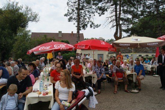 Eine Gruppe von Menschen sitzt unter Schirmen an Tischen in einem Outdoor-Essbereich. Einige tragen Brillen, und es gibt Flaschen und Essen auf den Tischen. Bäume und Gebäude sind im Hintergrund.
