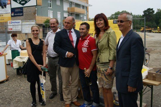 Eine Gruppe von Menschen, darunter ein Mann mit einer Flasche, steht vor einer Baustelle mit einem gelben Banner und einem im Bau befindlichen Gebäude im Hintergrund.