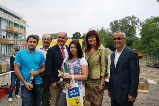 Eine Gruppe von Menschen steht draußen zusammen, einer hält eine Flasche Champagner, ein anderer ein Kuscheltier und eine Frau hält eine Tasche. Hinter ihnen steht ein Gebäude und einige Bäume.