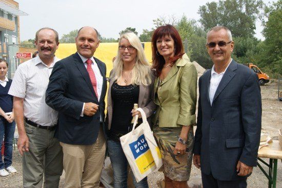 Fünf Personen stehen zusammen vor einer Baustelle. Die Frau in der Mitte hält eine weiße Tasche mit blauem und gelbem Text. Der Mann links trägt ein gestreiftes Hemd und einen Gürtel. Der Mann rechts trägt einen Anzug und eine Sonnenbrille. Hinter ihnen befindet sich eine gelbe Wand und ein Gebäude.