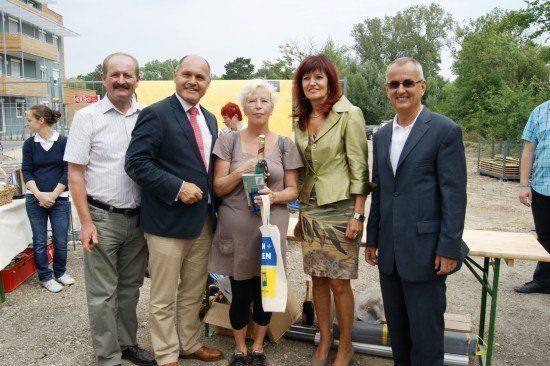 Eine Gruppe von Menschen, wahrscheinlich bei einer Outdoor-Veranstaltung, steht zusammen mit Lächeln auf den Gesichtern. Die Frau in der Mitte hält eine Flasche und eine Tasche. Dahinter steht ein Tisch mit einigen Gegenständen darauf.