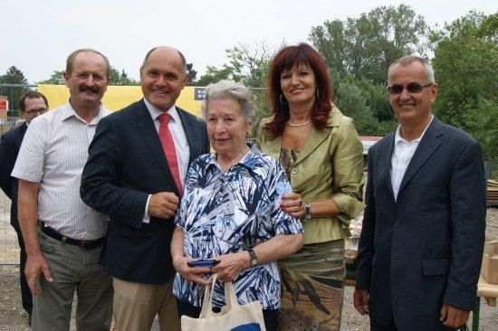 Fünf Menschen stehen zusammen in einer Gruppe und lächeln für ein Foto. Sie sind in formeller Kleidung gekleidet, und eine Frau hält eine Handtasche. Hinter ihnen befindet sich eine Baustelle mit Bäumen und einem gelben Banner.