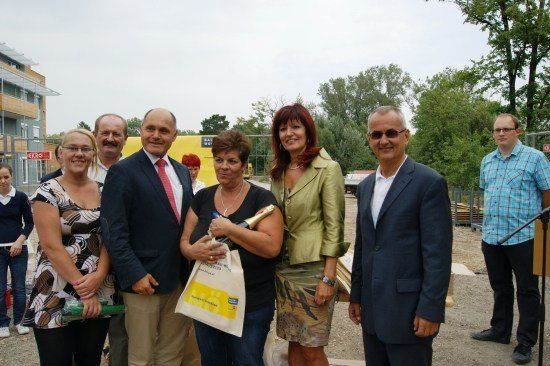 Eine Gruppe von Menschen in formeller Kleidung steht draußen an einem sonnigen Tag, lächelt und posiert für ein Foto. Eine Frau hält eine Flasche und eine Tasche. Hinter ihnen befindet sich eine Baustelle mit einer gelben Struktur und Bäumen.