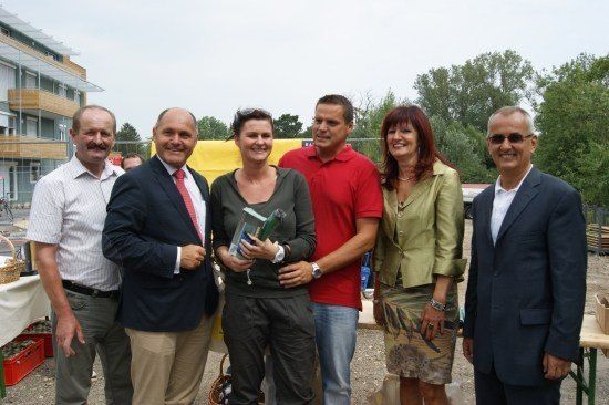 Eine Gruppe von Menschen in formeller Kleidung steht lächelnd für ein Foto zusammen. Eine Frau hält eine Trophäe. Hinter ihnen sind ein Gebäude und Bäume zu sehen.