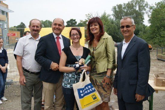 Eine Gruppe von Menschen steht zusammen, lächelt und posiert für ein Foto. Eine Frau in der Mitte hält ein Plüschtier und eine Tasche. Hinter ihnen befindet sich eine gelbe Wand und ein Zaun.