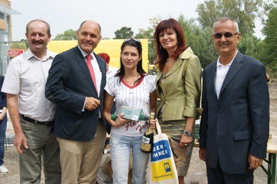 Fünf Menschen stehen lächelnd in einer Gruppe. Eine Frau hält eine Champagnerflasche und eine Geschenkbox. Im Hintergrund ist eine Baustelle mit Bäumen zu sehen.
