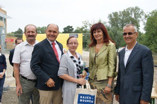 Eine Gruppe von Menschen in formeller Kleidung steht vor einer Baustelle und lächelt für die Kamera. Eine Frau hält eine Tasche mit dem Text 'Bauen Wohnen'. Hinter ihnen ist eine gelbe Wand mit einem Schild und Bäumen zu sehen.