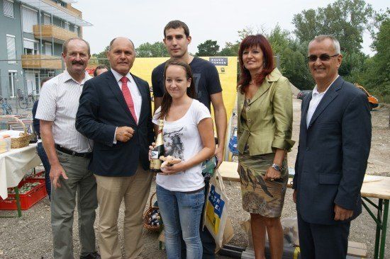 Eine Gruppe von Menschen steht zusammen, lächelt und posiert für ein Foto. Eine Frau in der Mitte hält eine Flasche Champagner. Hinter ihnen steht ein Tisch mit einem Korb und einem gelben Banner.