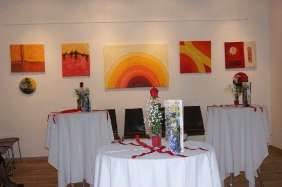 Ein Kunstgalerie mit drei Gemälden an der Wand und drei Tischen mit Blumen und Bändern.