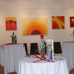 Ein Kunstgalerie mit drei Gemälden an der Wand und drei Tischen mit Blumen und Bändern.