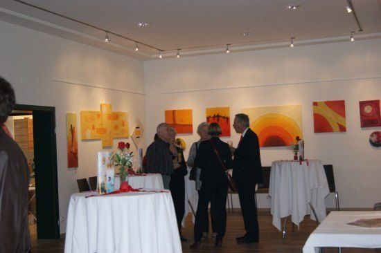 Eine Gruppe von Menschen steht in einer Kunstgalerie und diskutiert die Gemälde an den Wänden. Der Raum ist gut beleuchtet und die Gemälde sind an weißen Wänden ausgestellt.