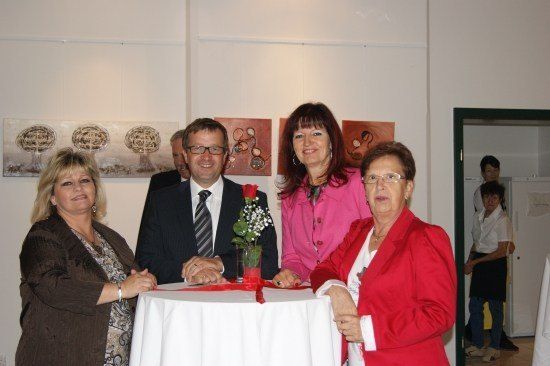 Vier Erwachsene stehen um einen Tisch mit Blumenarrangement und lächeln für ein Foto in einem Raum mit weißen Wänden und gerahmten Kunstwerken.
