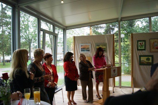 Eine Gruppe von Frauen in roten Jacken steht vor einem Podium mit einem Mikrofon. Sie befinden sich in einem Raum mit großen Fenstern und ausgestellten Gemälden.
