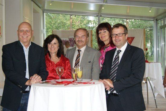 Fünf Personen, drei Männer und zwei Frauen, stehen um einen Tisch mit Getränken und lächeln für ein Foto. Die Frau in Rot hält die Hand des Mannes auf ihrer linken Seite.