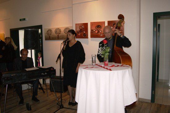 Eine Frau steht an einem Mikrofon und singt, während ein Mann Cello spielt. Ein Pianist sitzt hinter ihr, und ein Tisch mit Blumenarrangement steht vor ihnen.