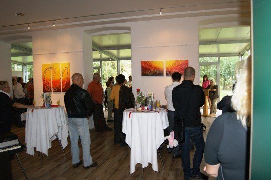 Eine Gruppe von Menschen versammelt sich in einem Innenraum, möglicherweise einer Kunstgalerie, mit Tischen und Gemälden an der Wand.