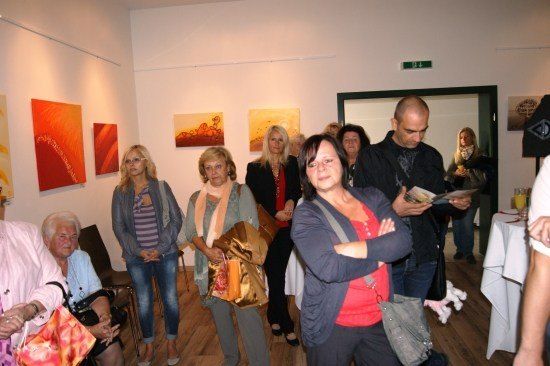 Eine Gruppe von Menschen versammelt sich in einer Kunstgalerie, einige betrachten Gemälde an der Wand, während andere sprechen.