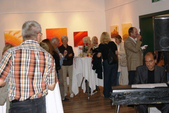 Eine Gruppe von Menschen versammelt sich in einem Innenraum, möglicherweise einer Kunstgalerie, wobei einige um einen Tisch mit Getränken stehen und andere sich unterhalten. Ein Mann spielt in der Ecke Klavier.