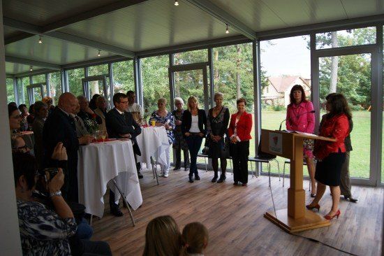Eine Gruppe von Menschen ist in einem Raum mit Glaswänden versammelt, wahrscheinlich für eine Veranstaltung. Eine Frau spricht an einem Podium, während andere hinter ihr stehen. Tische mit weißen Tischdecken sind aufgestellt, und einige Leute sitzen. Draußen gibt es einen Rasenbereich mit Bäumen.