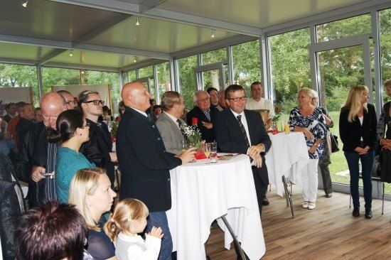 Eine Gruppe von Menschen in formeller Kleidung versammelt sich in einem Raum mit großen Fenstern, einige stehen, andere sitzen. Ein Mann in Anzug hält ein Glas und spricht mit einem anderen Mann. Es gibt Tische mit weißen Tischdecken und Blumen. Draußen gibt es Bäume und ein Grasfeld.