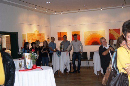 Eine Gruppe von Menschen versammelt sich in einer Kunstgalerie, steht um Tische mit Blumen und Getränken herum, während andere Gemälde an der Wand betrachten.