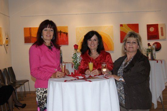 Drei Frauen stehen um einen Tisch in einer Kunstgalerie herum, halten Gläser mit Orangensaft und lächeln. Hinter ihnen sind mehrere Gemälde an der Wand ausgestellt.