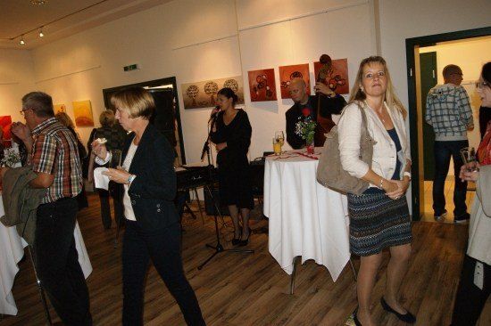 Eine Gruppe von Menschen ist in einem Innenraum versammelt, möglicherweise in einer Kunstgalerie, mit einer Frau, die in ein Mikrofon singt, und einem Mann, der Cello spielt.