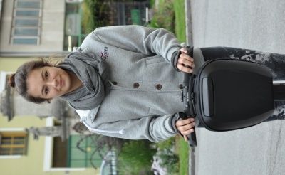 Eine Frau in einer grauen Kapuzenjacke und einem Schal hält einen schwarzen Elektroroller auf einer Straße.