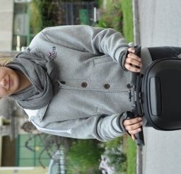 Eine Frau in einer grauen Kapuzenjacke und einem Schal hält einen schwarzen Elektroroller auf einer Straße.