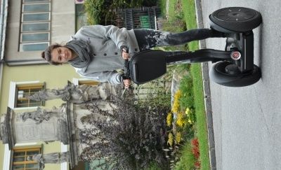 Eine Frau fährt auf einem schwarzen Segway auf einer Straße, mit einer Statue und Gebäuden im Hintergrund.